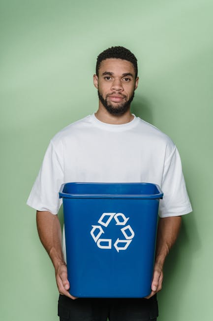 A young man with a neutral facial expression, short curly dark hair, and a beard, standing against a pale green background, holding a large blue recycling bin. The bin features a white universal recyc
