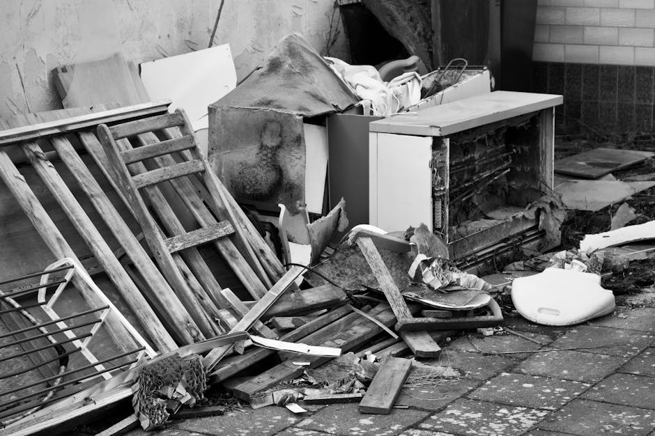 A collection of discarded household items and debris arranged outside on a paved surface, featuring a wooden garden chair with slatted seating and backrest leaning against a pile of rubbish, and a par